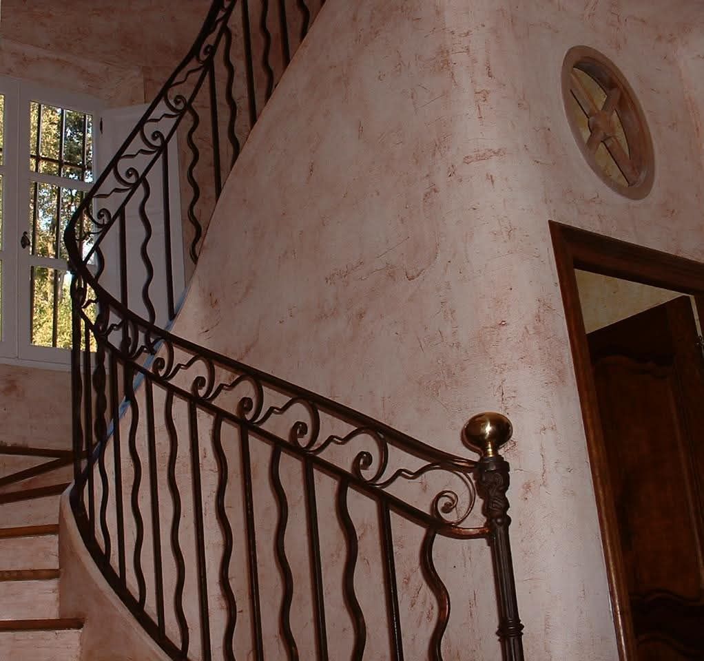 Un escalier courbe avec une rampe en métal sombre ornée se détache sur un mur texturé dans un couloir intérieur.