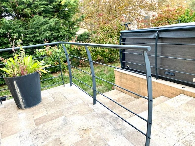 Une rambarde en métal épare une terrasse pavée ornée d'une grande plante en pot d'un jacuzzi et d'un escalier dans un jardin.