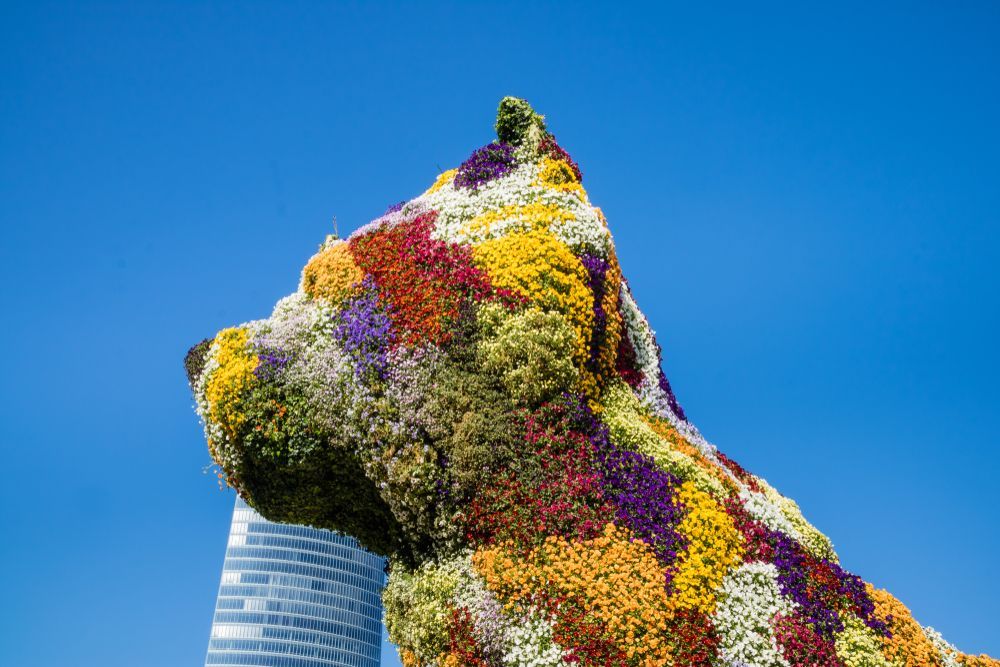 Gran escultura floral de perro con flores de colores contra un cielo azul y un edificio alto.