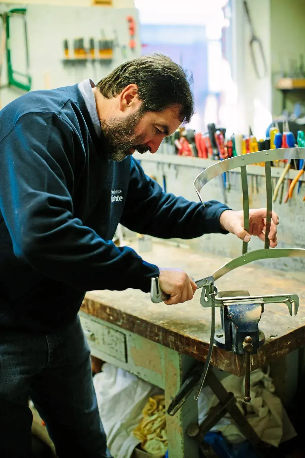 Ein Mann bearbeitet an einer Werkbank in einer Werkstatt einen Metallgegenstand. Er benutzt ein Werkzeug.
