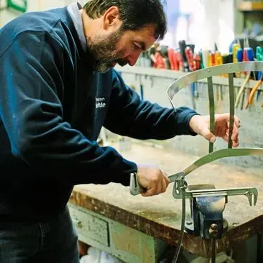 Eine Person arbeitet in einer Werkstatt an Metallstuhlteilen und benutzt dabei ein Werkzeug.