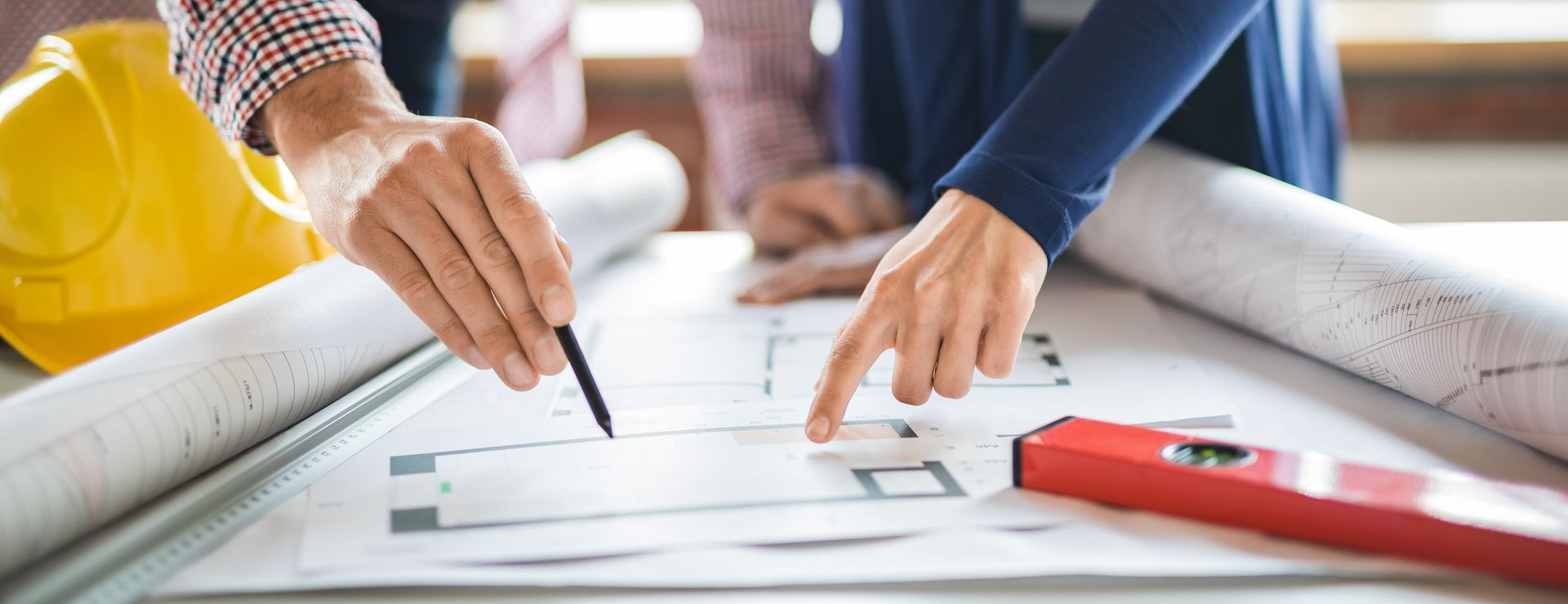 Deux personnes examinent des plans de construction sur une table, avec un casque de chantier jaune et un niveau rouge à proximité.