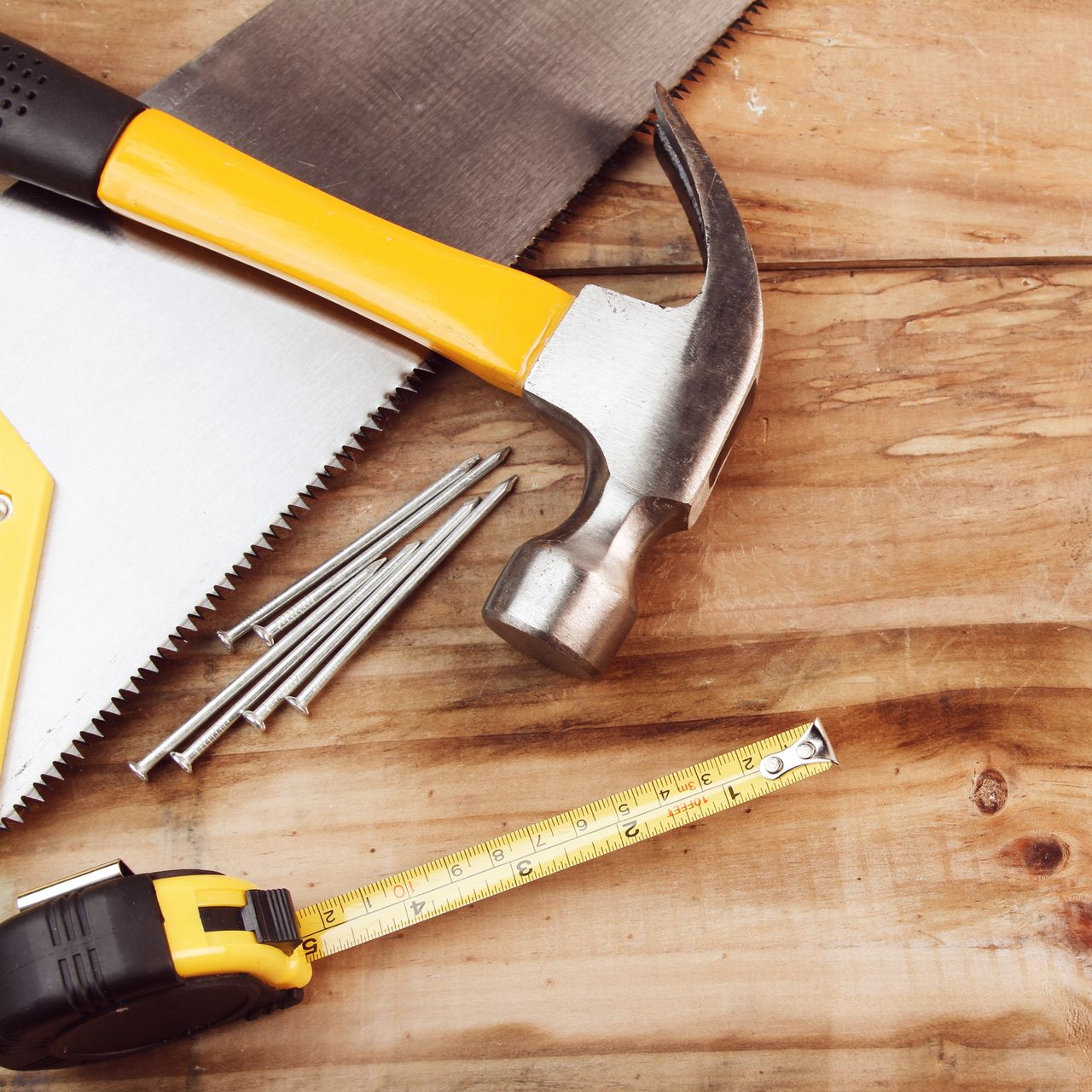 Un marteau à manche jaune, une scie à main, un mètre ruban et plusieurs clous disposés sur une surface en bois.