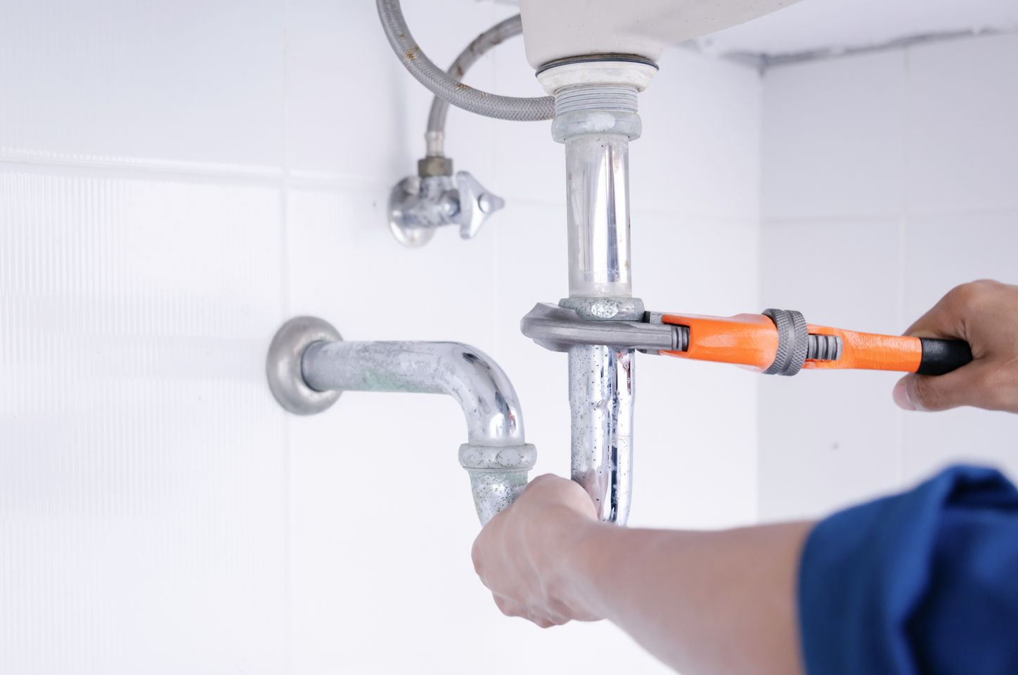 Une personne utilise une clé à pipe orange pour serrer un tuyau métallique sous un lavabo contre un mur carrelé blanc.