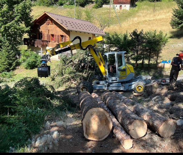 Une excavatrice jaune travaille sur un tas de bûches devant une maison