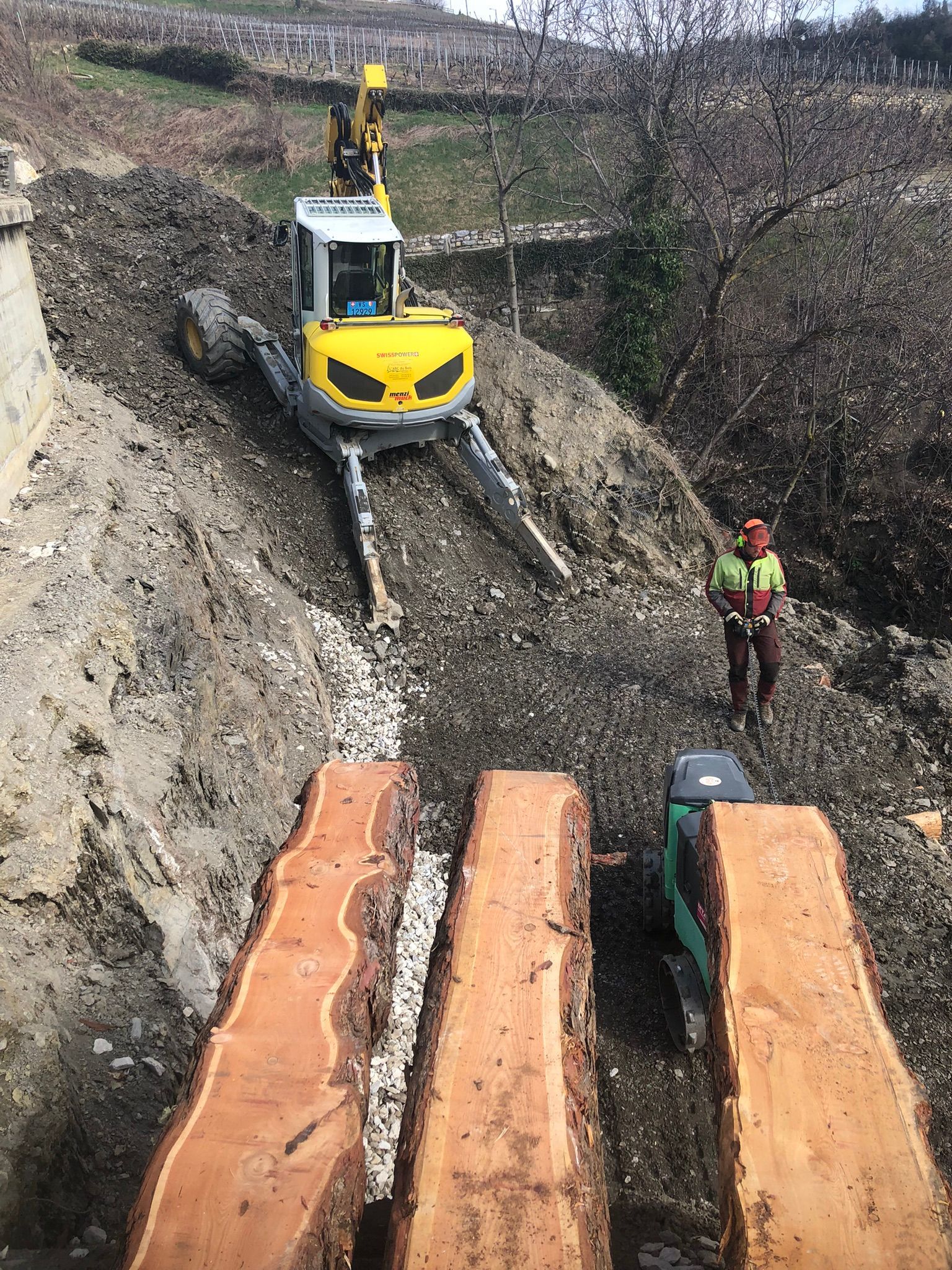 Une excavatrice jaune roule sur un chemin de terre à côté d'un tas de bûches.
