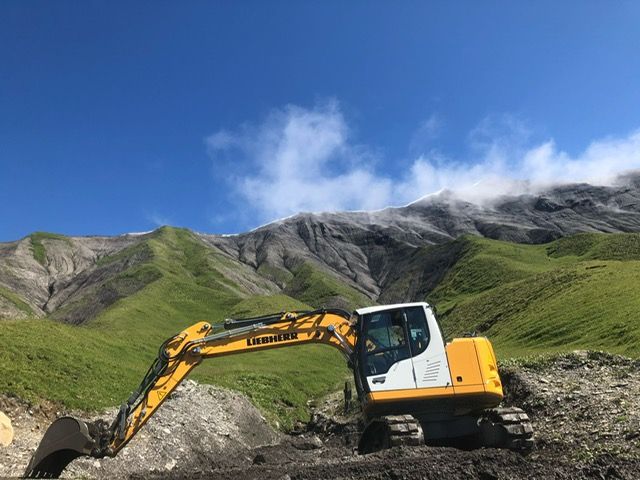 Une excavatrice jaune roule sur un chemin de terre dans les montagnes.