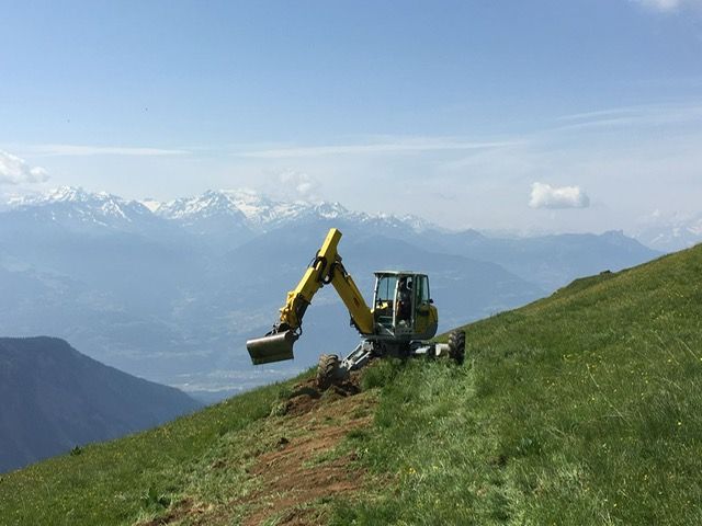 Une excavatrice jaune est assise au sommet d'une colline herbeuse avec des montagnes en arrière-plan.