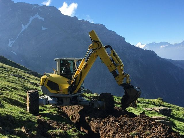 Une excavatrice jaune creuse un trou dans un champ herbeux avec des montagnes en arrière-plan