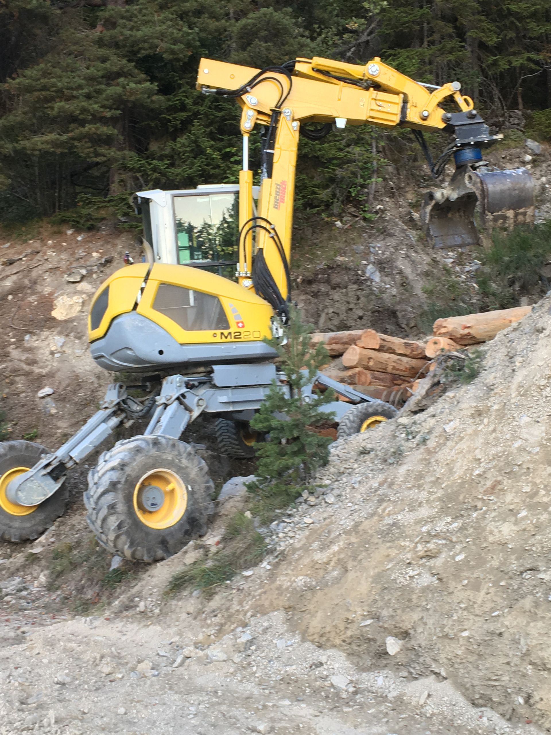 Une excavatrice jaune est assise au sommet d'une colline de terre à côté d'un tas de bûches.