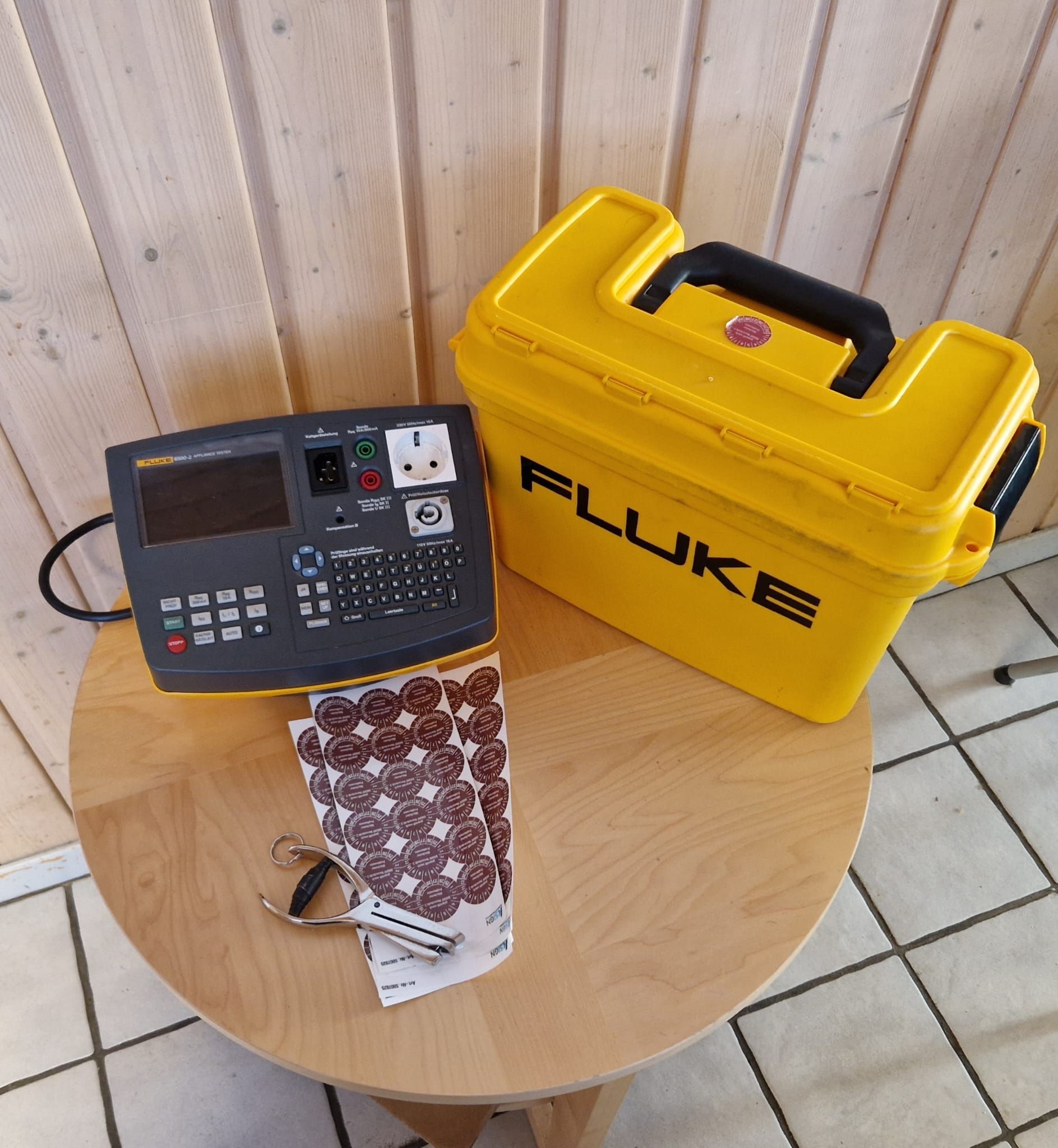 Elektrischer Tester von Fluke und gelbes Gehäuse auf einem Holztisch. Daneben liegen ein Bogen mit Aufklebern und ein Schlüsselanhänger.