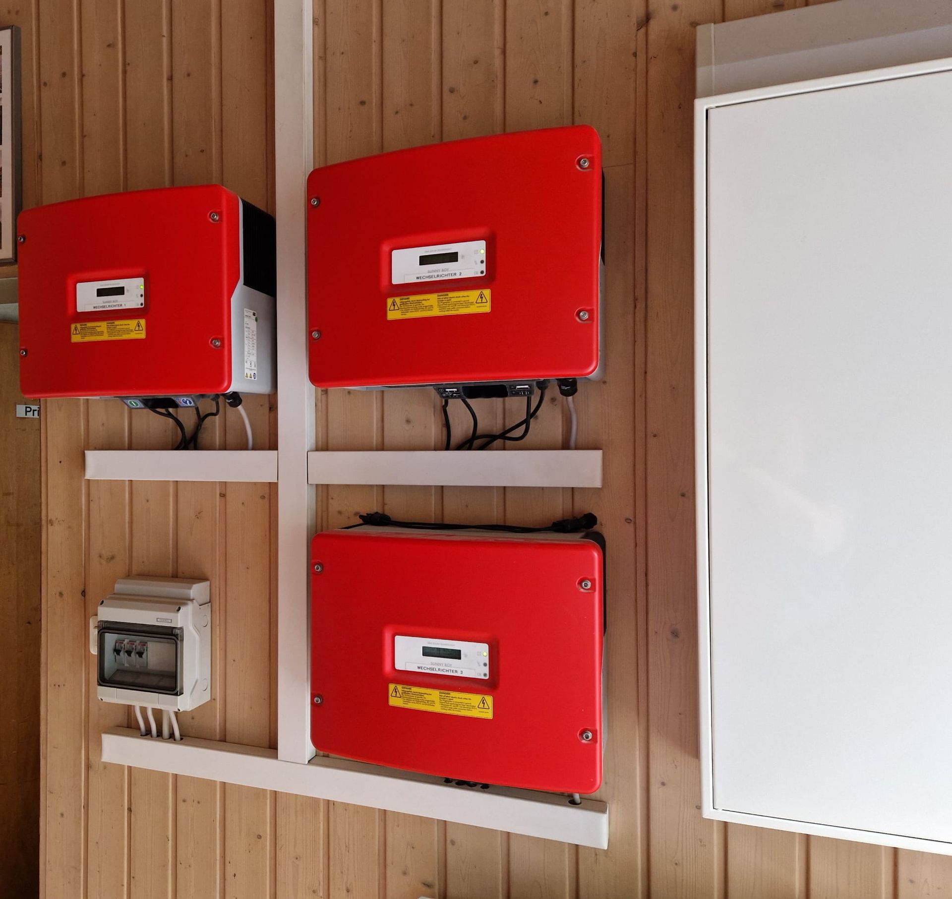 Drei rote Solarwechselrichter sind an einer Holzwand montiert; außerdem sind ein weißes Schaltfeld und ein weißer Kasten zu sehen.