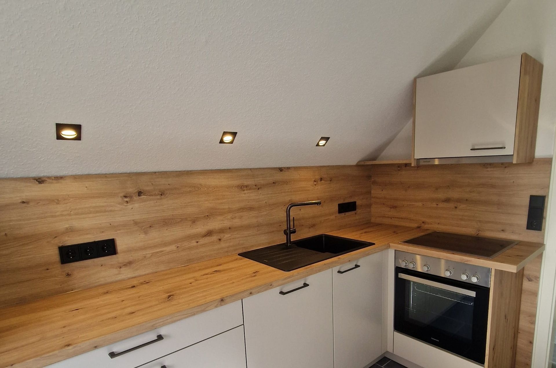 Kleine Küche mit Küchenrückwand in Holzoptik, weißen Schränken, schwarzer Spüle und Wasserhahn, Backofen und Einbauleuchten.
