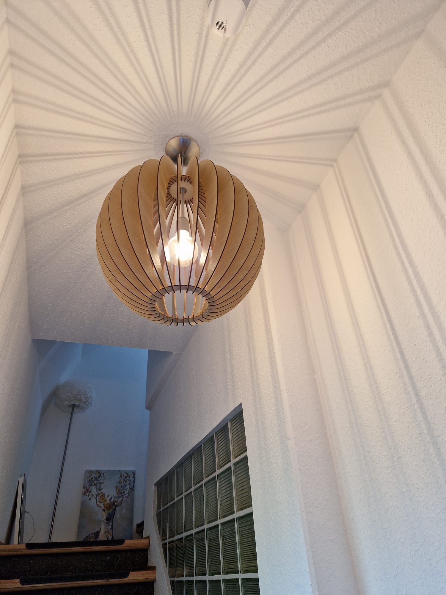 Kugelförmige Pendelleuchte aus Holz, die radiale Schatten auf die weiße Treppenhausdecke wirft.