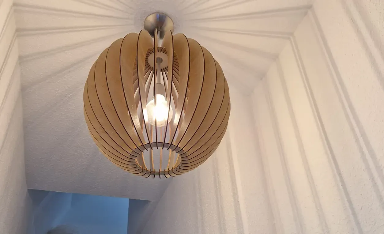 In einem weißen Flur hängt eine hölzerne Pendelleuchte in Kugelform, die einen sternförmigen Schatten wirft.