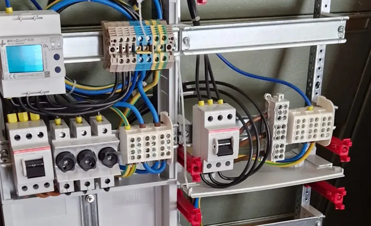 Schalttafel mit verschiedenen Leistungsschaltern, Kabeln und einem Messgerät. Blaue, gelbe und schwarze Kabel sind sichtbar.