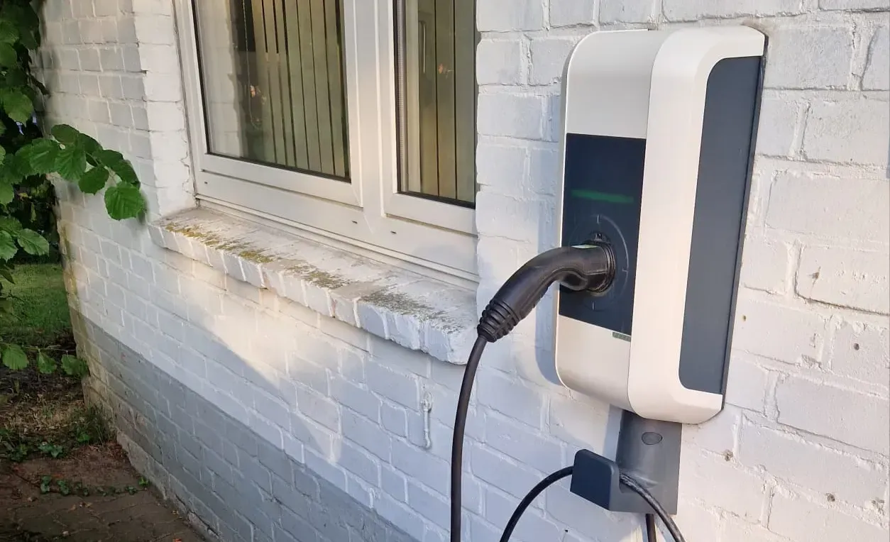 Ladegerät für Elektrofahrzeuge an einer weißen Ziegelwand neben einem Fenster montiert, Kabel eingesteckt.