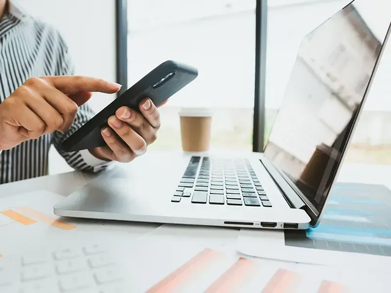 Person, die in einem hellen Büro ein Smartphone neben einem Laptop und einer Kaffeetasse verwendet.