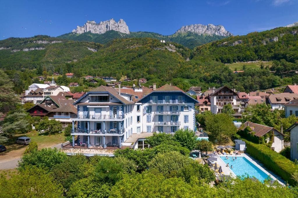 Station de montagne avec bâtiments blancs, toits bleus, piscine et pics rocheux sous un ciel dégagé.