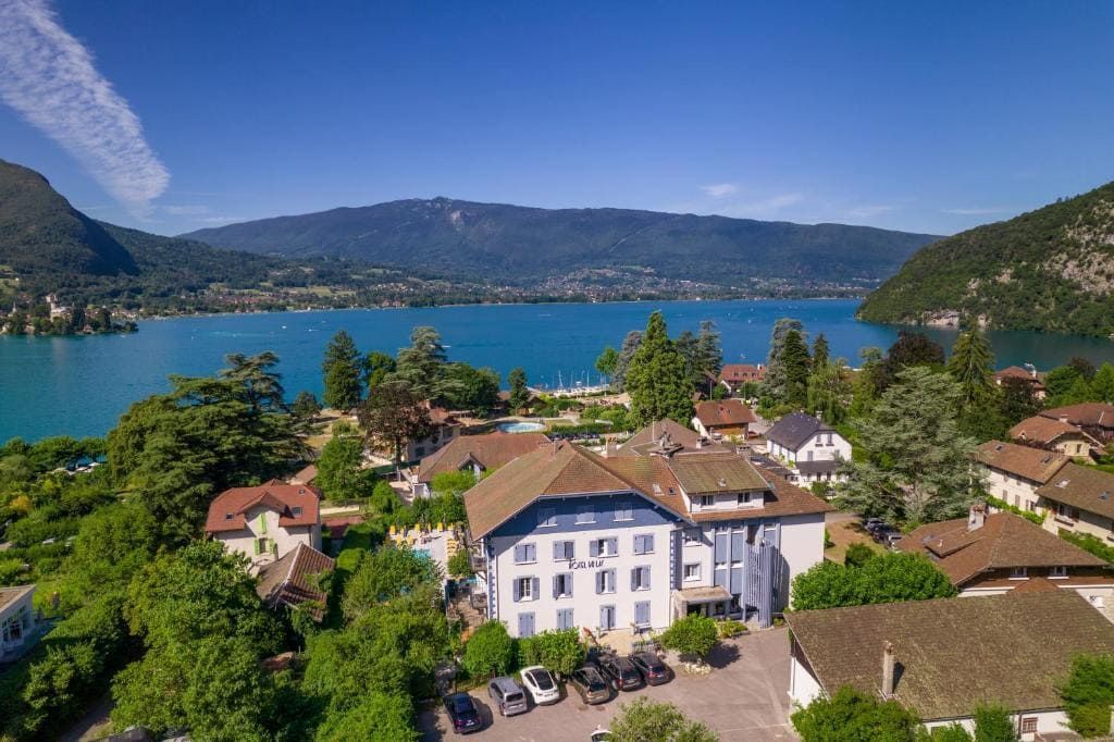 Village au bord d'un lac avec des bâtiments blancs, des toits rouges et des montagnes sous un ciel bleu clair