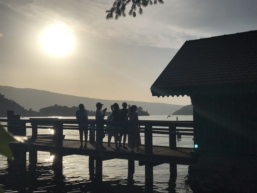 Des silhouettes de personnes se tiennent sur un quai au coucher du soleil, sur une eau calme, avec des montagnes en arrière-plan.