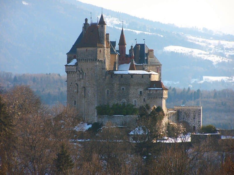 Château de pierre aux hautes tours sur un flanc de colline enneigé, encadré par des arbres dénudés et des montagnes brumeuses