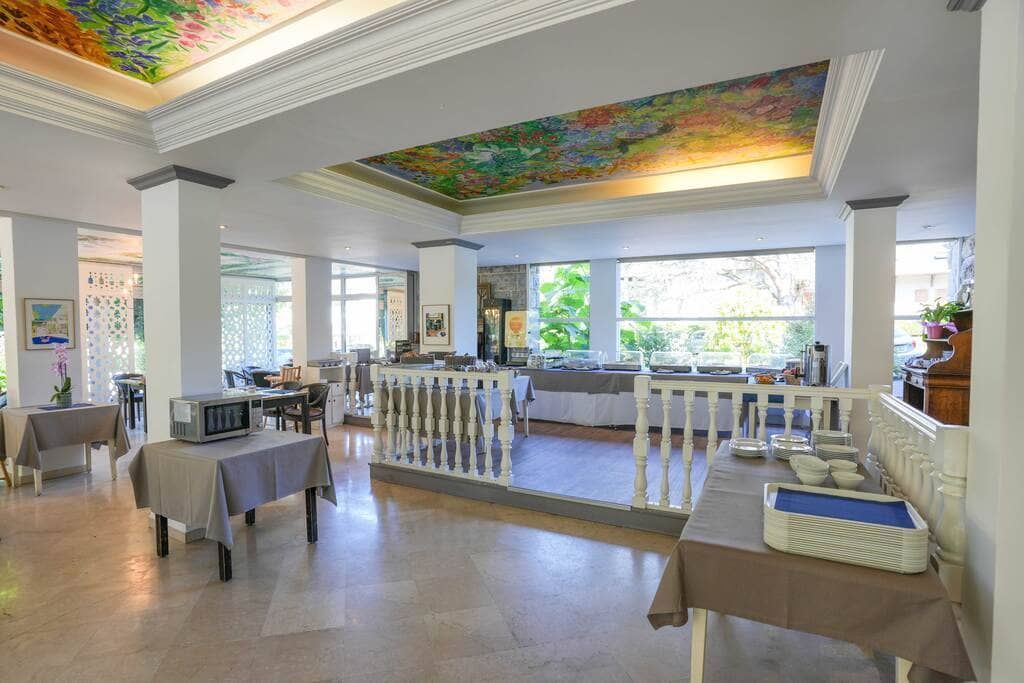 Salle à manger lumineuse d'un restaurant avec tables et chaises blanches et une fresque colorée au plafond.