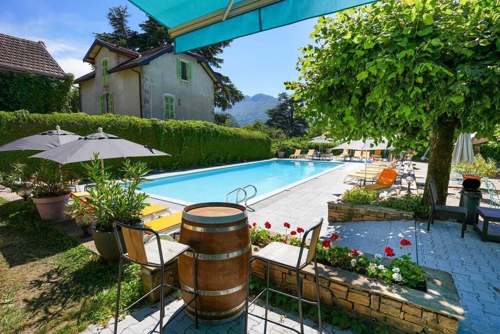 Piscine ensoleillée dans le jardin avec chaises longues blanches, haies vertes et patio ombragé à côté d'une maison rustique