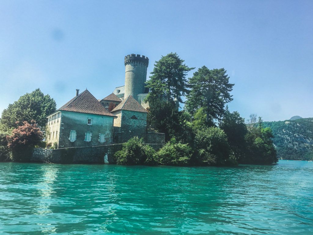 Petit château de pierre avec une tour, situé sur un rivage luxuriant au bord d'une eau turquoise éclatante.