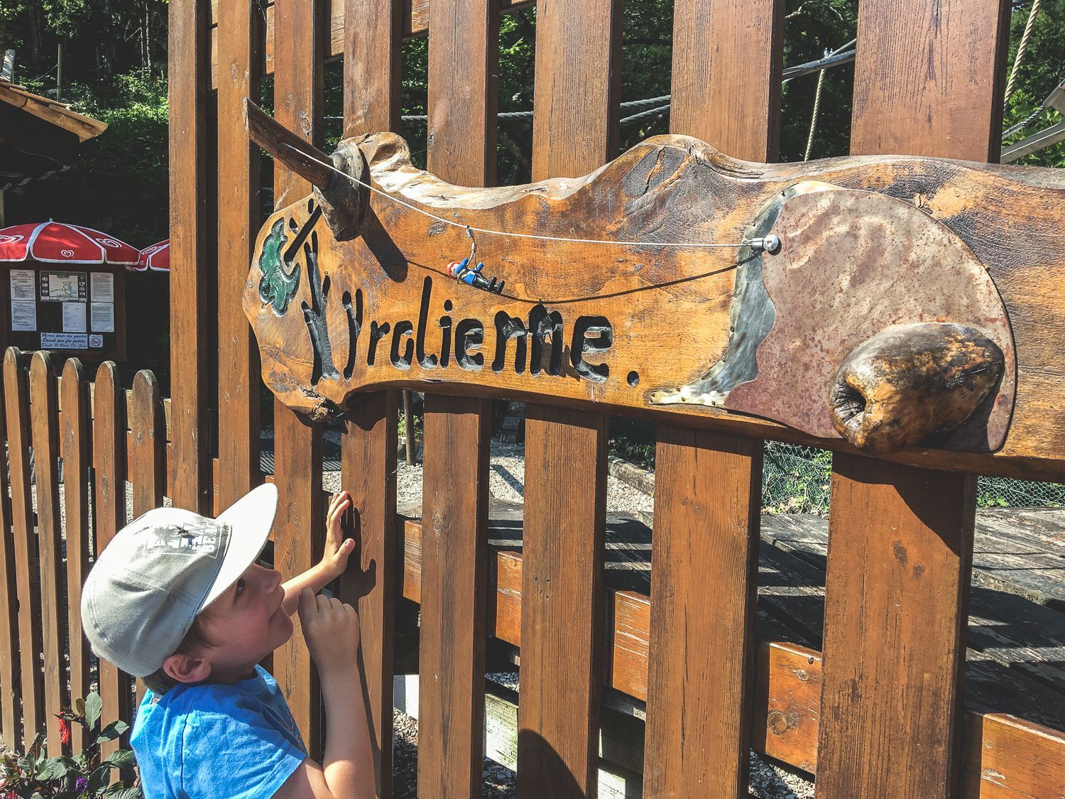 Un enfant en chemise bleue et casquette montre du doigt un panneau en bois représentant un animal, près d'une clôture.