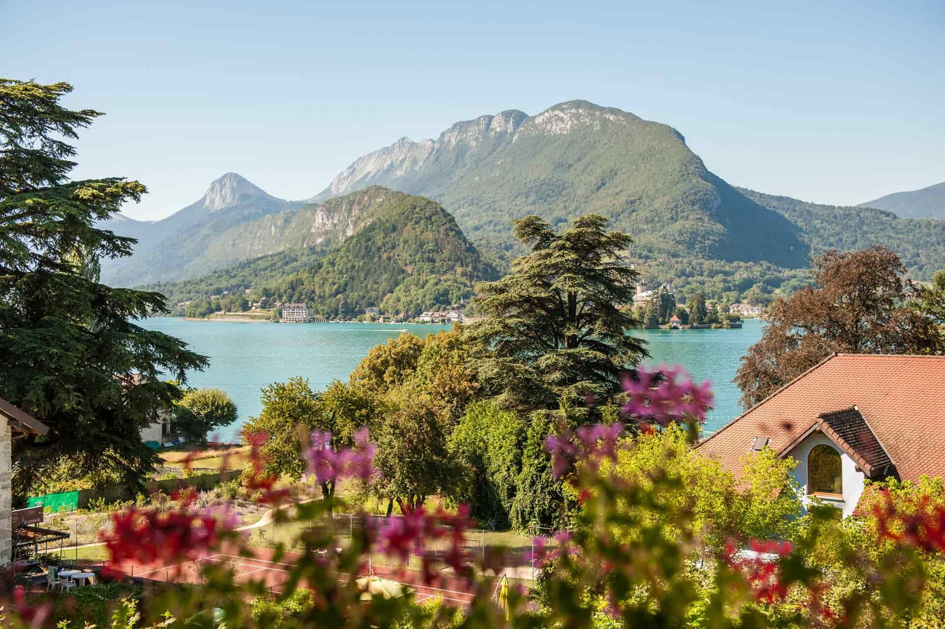 Vue pittoresque sur un lac et des montagnes, avec des fleurs colorées, des arbres et un chalet au toit rouge au premier plan.