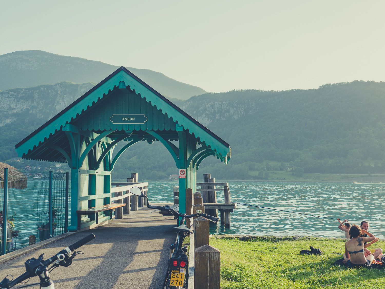 Un kiosque à toit turquoise sur une jetée au bord d'un lac, avec des montagnes en arrière-plan et des personnes assises sur l'herbe.