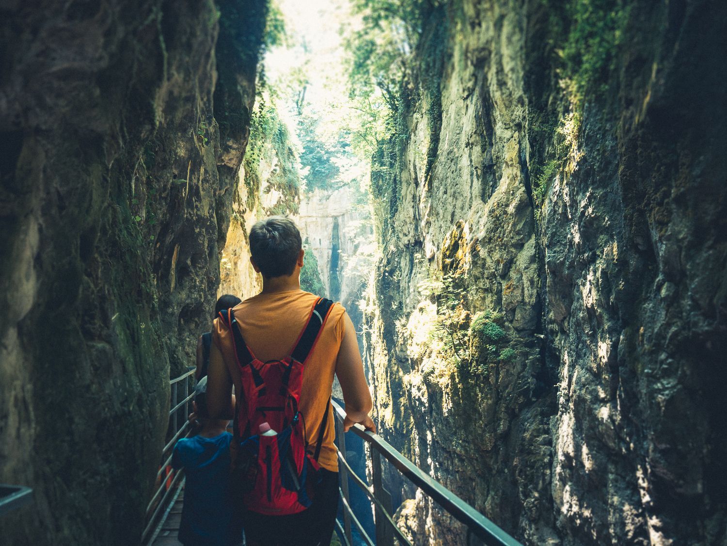 Randonneur avec un sac à dos rouge marchant sur une passerelle métallique dans une gorge rocheuse étroite