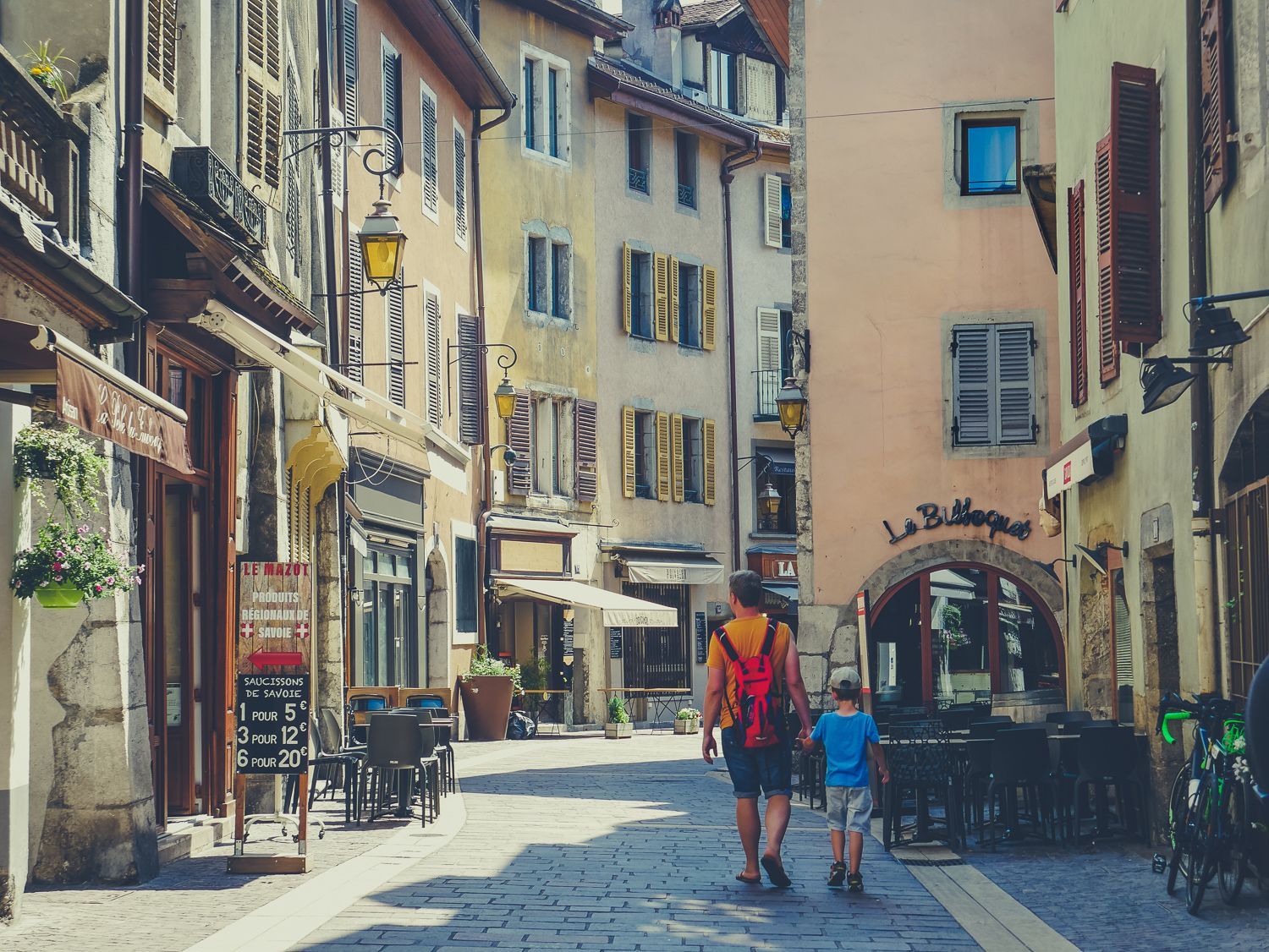 Rue européenne étroite bordée de bâtiments colorés, de cafés en terrasse et de deux personnes marchant sous une arche ensoleillée.