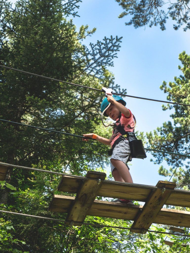 Une personne en chemise rose et harnais traverse un pont de corde dans la canopée d'une forêt feuillue.