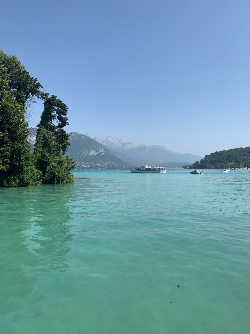 Lac turquoise avec un bateau près d'une rive boisée et des montagnes brumeuses sous un ciel bleu clair