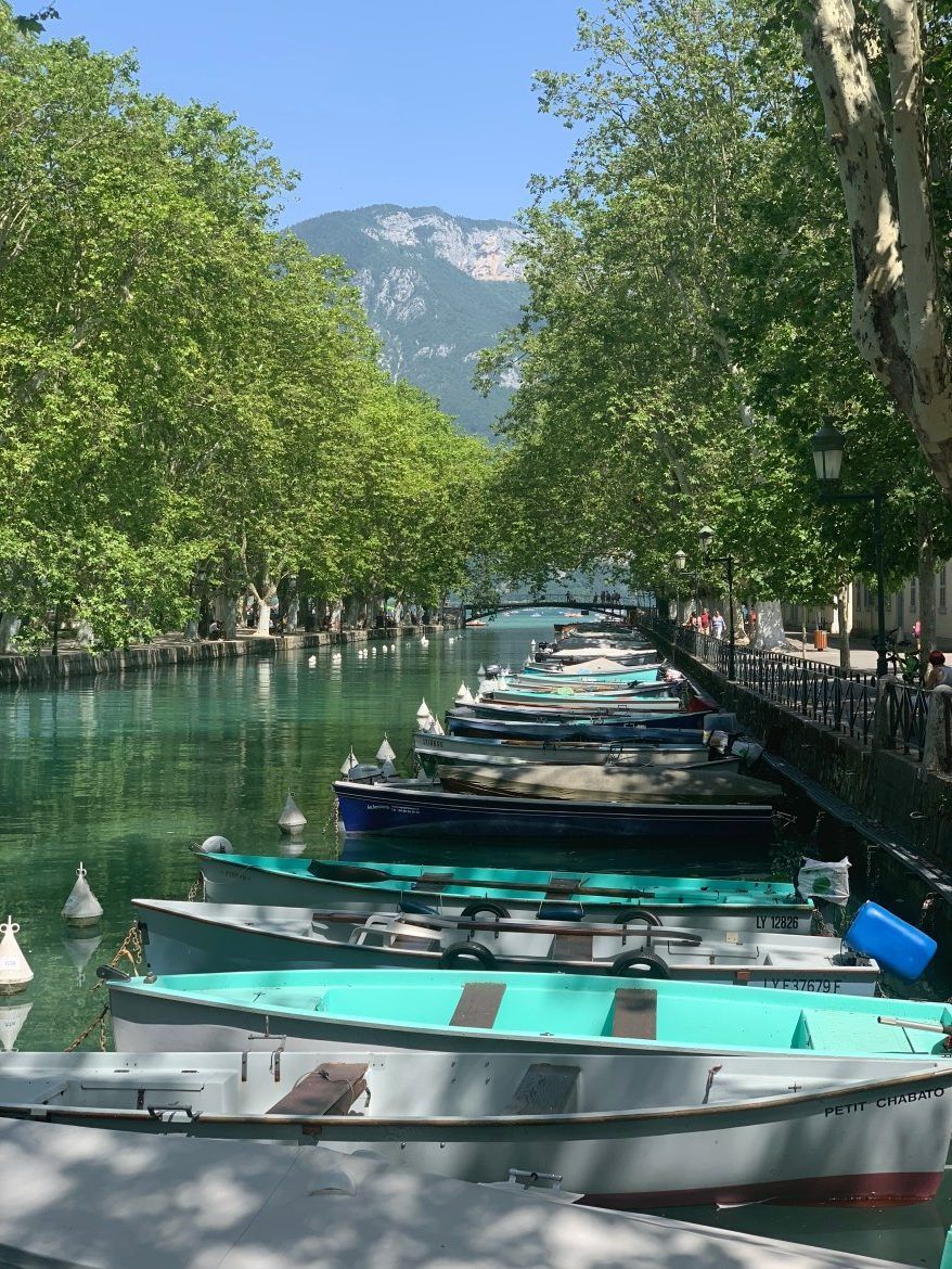 Une rangée de bateaux amarrés sur un canal turquoise bordé d'arbres, avec des montagnes à l'horizon.