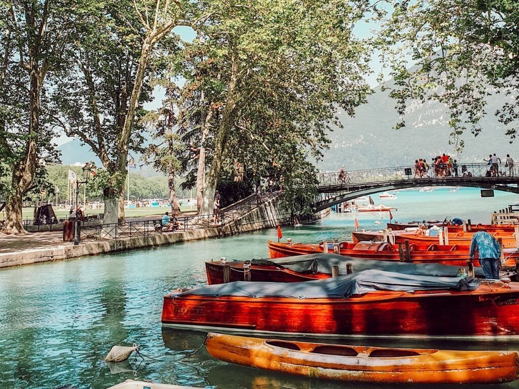 Des bateaux rouges et oranges naviguent sur un canal bleu, bordé d'arbres et d'un pont piétonnier.