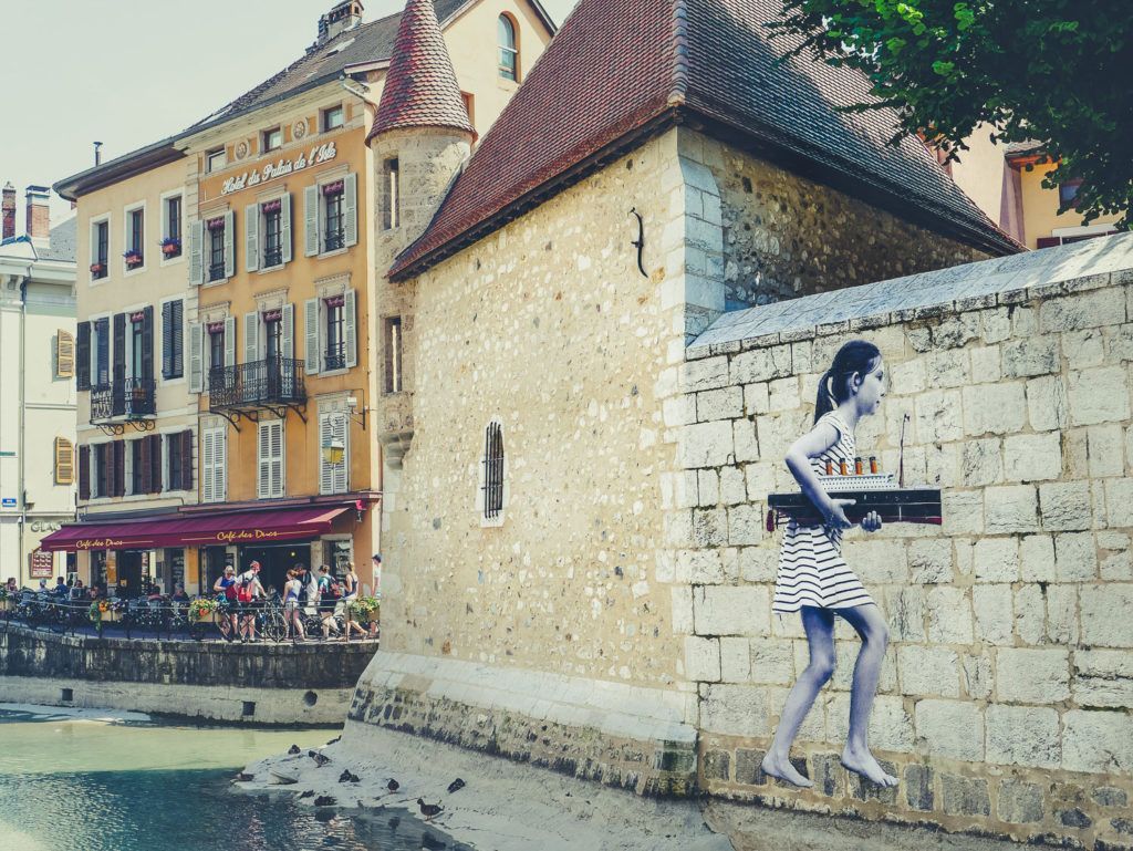 Une fresque murale représentant une personne sur un mur de pierre au bord d'un canal, avec des bâtiments colorés et des terrasses de café en arrière-plan.