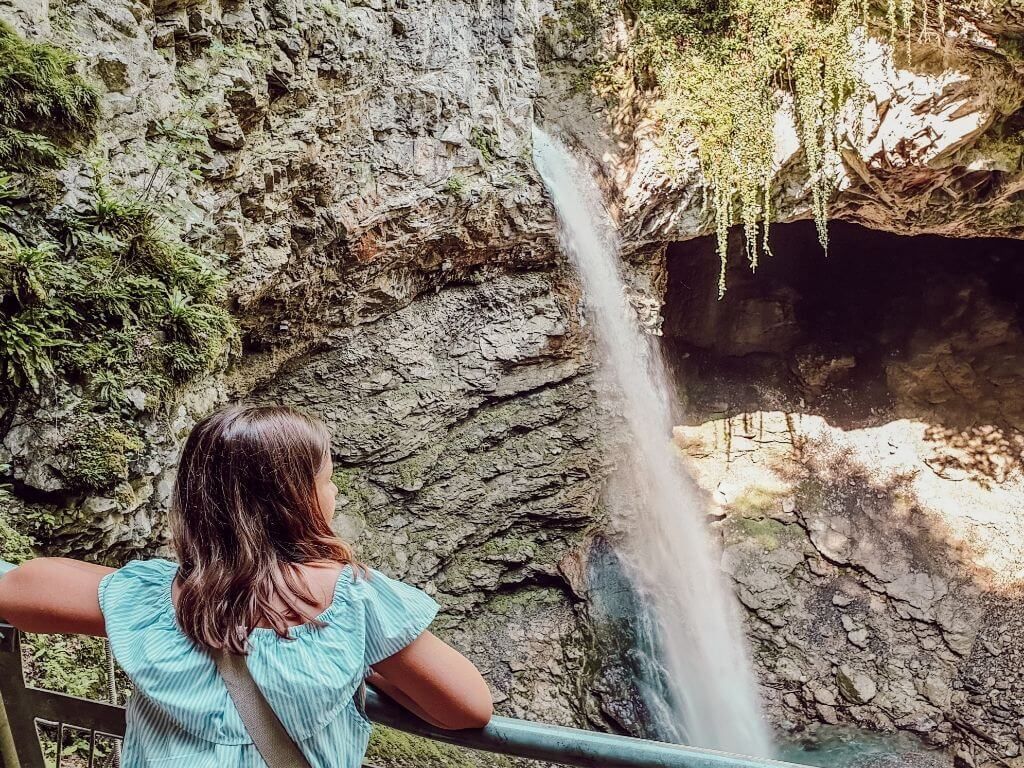 Une personne vêtue d'une chemise turquoise se tient près d'une cascade dans une grotte rocheuse.