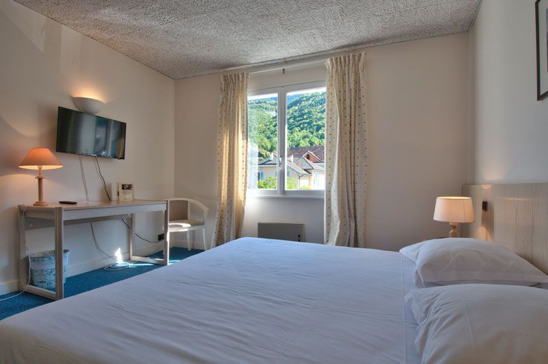 Chambre lumineuse avec un lit, un bureau, des lampes et une fenêtre donnant sur la verdure.