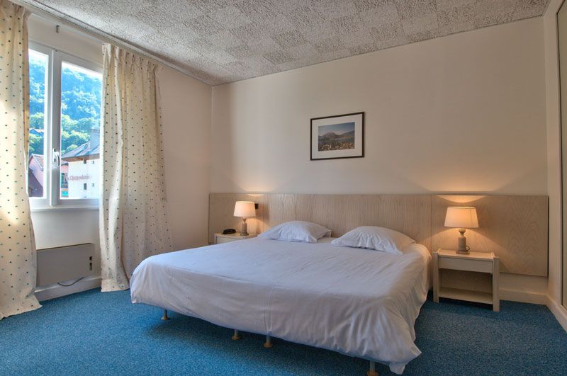 Chambre d'hôtel lumineuse avec un grand lit, des lampes de chevet, de la moquette bleue et une fenêtre avec des rideaux.