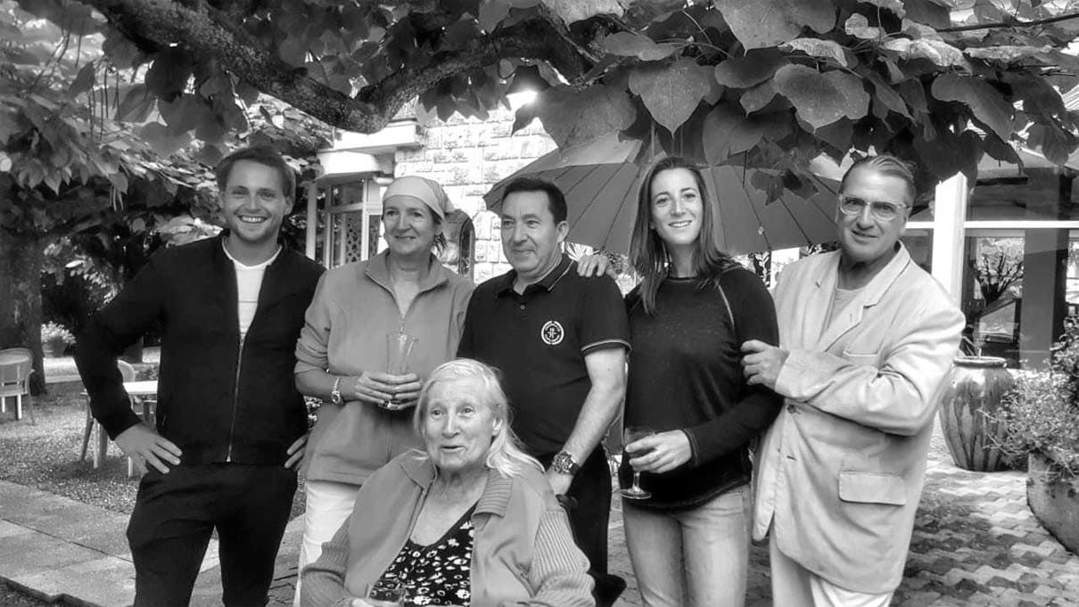 Un groupe de six personnes posant ensemble à l'extérieur, dans un jardin, souriantes et debout, une personne étant assise devant elles.