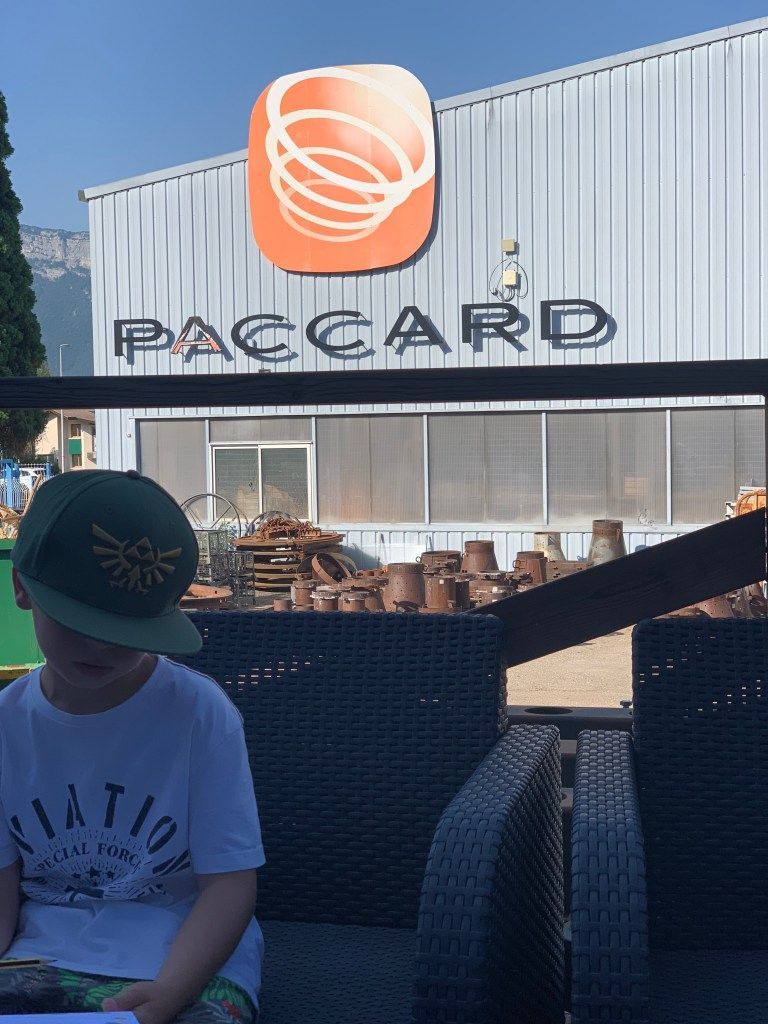 Terrasse extérieure devant un panneau PACCARD, avec une personne en chemise blanche et chapeau assise au premier plan.