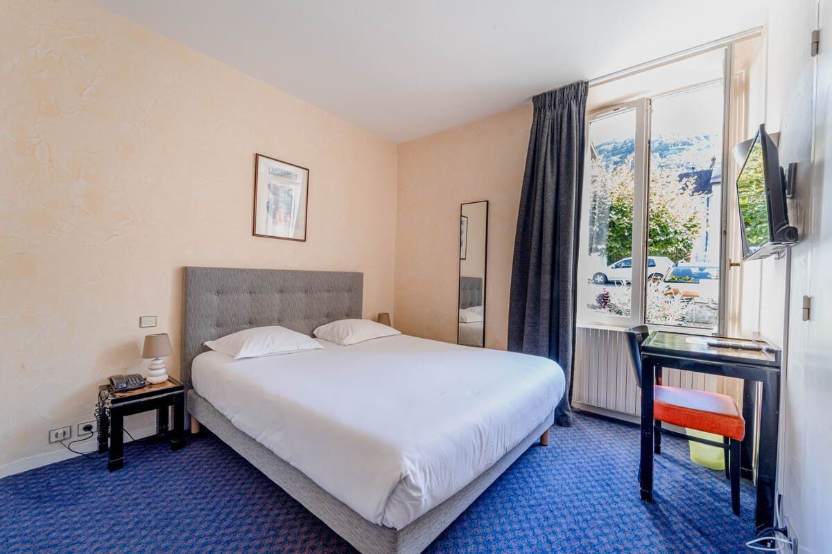 Chambre lumineuse avec un lit, une moquette bleue et une fenêtre ouverte laissant entrer la lumière du jour.