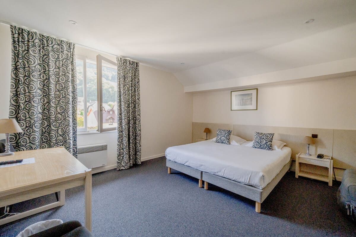 Chambre d'hôtel lumineuse avec deux lits, rideaux à motifs, une fenêtre et un petit bureau contre le mur.