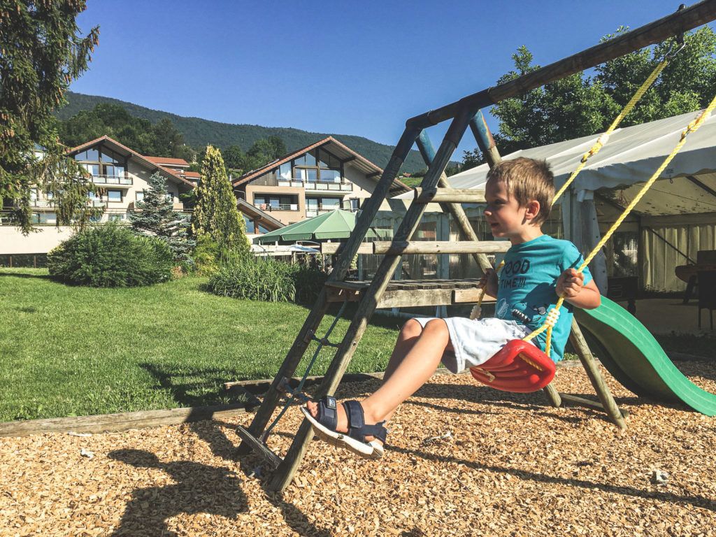 Enfant se balançant sur une balançoire dans un parc ensoleillé, avec des maisons et des montagnes en arrière-plan.