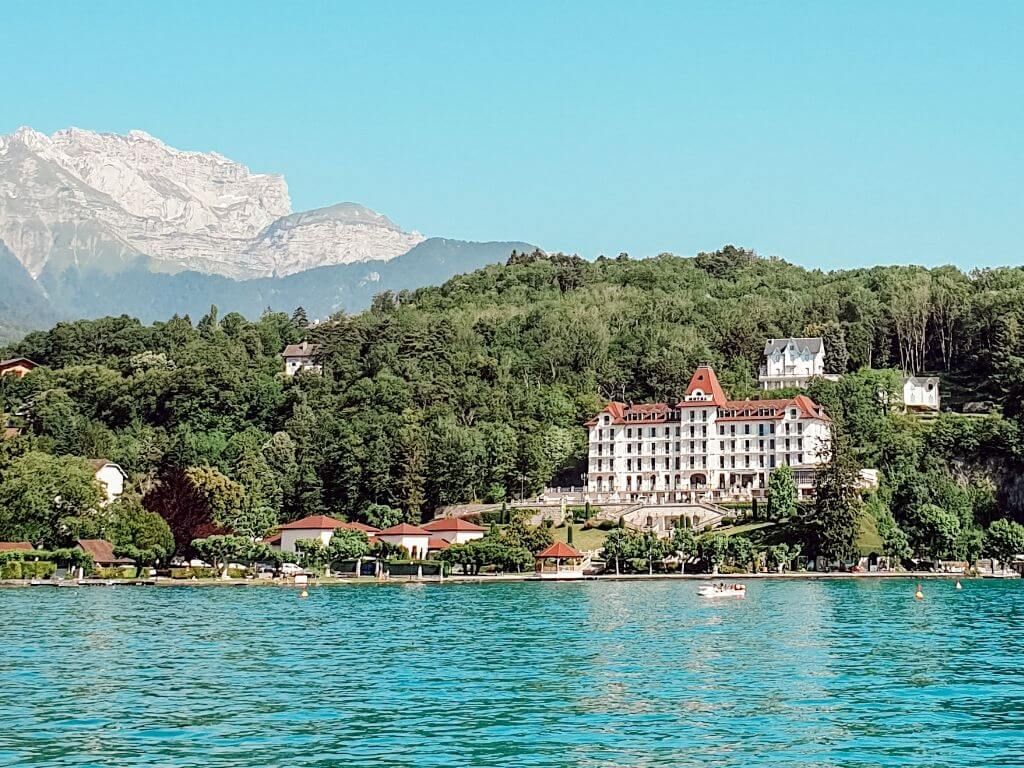 Complexe hôtelier au bord d'un lac avec un hôtel blanc, une eau turquoise, une rive boisée et des montagnes en arrière-plan.