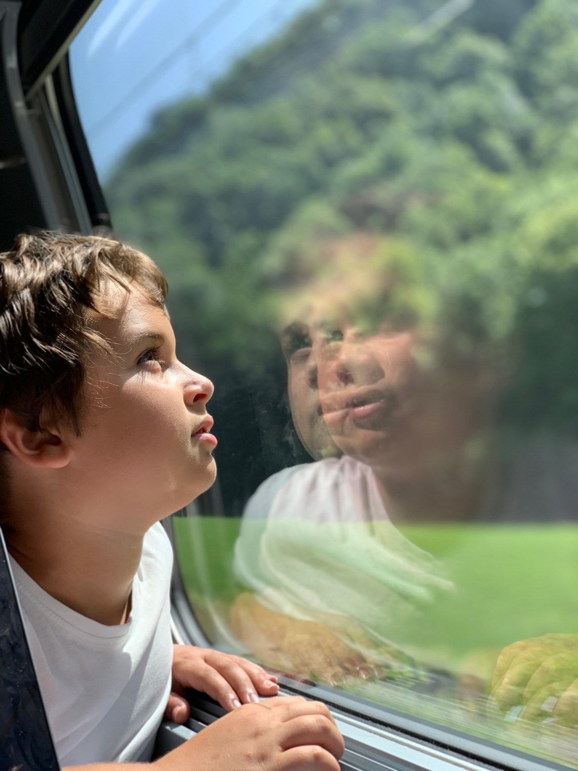 Un enfant regarde par la fenêtre d'un train, son visage se reflétant dans la vitre, avec les collines verdoyantes à l'horizon.