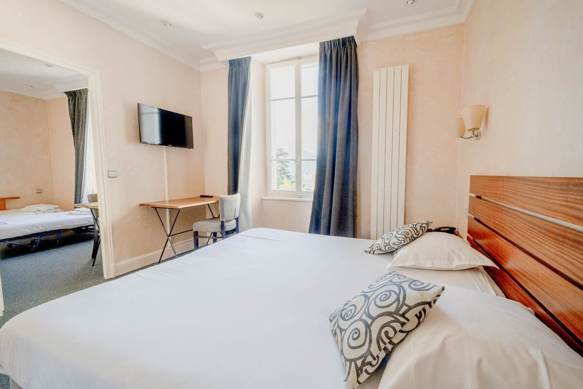 Chambre lumineuse avec un grand lit, des rideaux bleus, une télévision murale, un bureau et un miroir près de la porte.